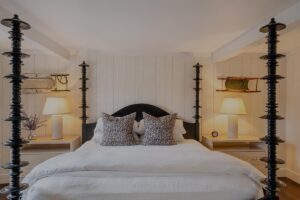 A cozy Edson Hill bedroom with a large bed, two patterned pillows, matching bedside tables, lamps, and shelves on a white paneled wall. Four dark decorative pillars frame the bed.