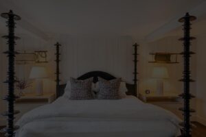 A cozy bedroom at Edson Hill features a four-poster bed, two patterned pillows, two bedside tables with lamps, and wooden shelves on the wall.