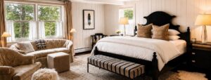 A cozy, elegant bedroom reminiscent of a Vermont Country Inn, with a large bed, striped bench, plush sofa, beige armchair, lamps, and two windows letting in natural light. Neutral tones and decorative pillows create a warm, inviting atmosphere.