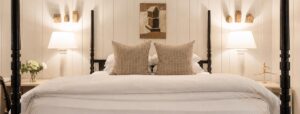 A neatly made bed with two patterned pillows, white bedding, and two bedside tables each holding a lamp creates a tranquil Edson Hill-inspired retreat. A flower vase and a small statue accent the tables, while geometric artwork on the wall above the bed completes this sophisticated setting.