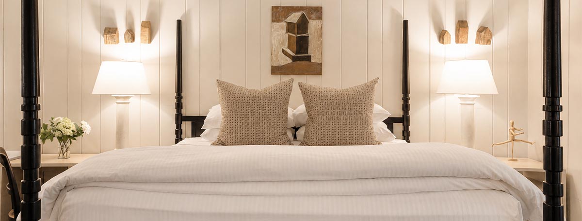 A neatly made bed with two patterned pillows, white bedding, and two bedside tables each holding a lamp creates a tranquil Edson Hill-inspired retreat. A flower vase and a small statue accent the tables, while geometric artwork on the wall above the bed completes this sophisticated setting. A neatly made bed with two patterned pillows, white bedding, and two bedside tables each holding a lamp creates a tranquil Edson Hill-inspired retreat. A flower vase and a small statue accent the tables, while geometric artwork on the wall above the bed completes this sophisticated setting.