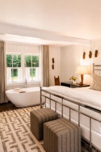 A cozy bedroom with a freestanding bathtub by large windows, a metal bed, patterned rug, striped ottomans, a desk with a lamp, and neutral decor evokes the timeless charm of Vermont luxury hotel accommodations. Sunlight streams through the curtains, enhancing the inviting ambiance.