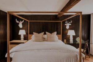 A cozy bedroom at Edson Hill features a wooden four-poster bed with white bedding, two beige pillows, two white lamps on nightstands, dark wood paneled walls, and two white decorative deer heads mounted above the lamps.