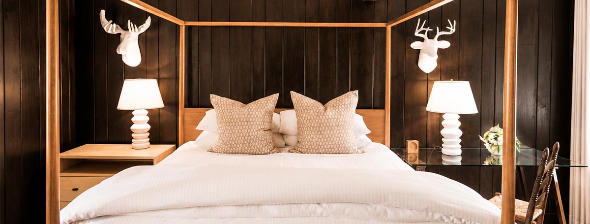 A cozy bedroom at Edson Hill features a wooden canopy bed, white bedding, two beige pillows, two white table lamps, light wood nightstands, and white decorative deer heads mounted on dark wood walls. A cozy bedroom at Edson Hill features a wooden canopy bed, white bedding, two beige pillows, two white table lamps, light wood nightstands, and white decorative deer heads mounted on dark wood walls.