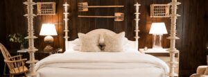 A cozy bedroom with a four-poster bed, white bedding, two lamps on nightstands, and wooden walls evokes the inviting charm of Edson Hill Guest Rooms. Decorative wooden tools hang above the bed, while woven baskets are mounted on each side, adding to the room’s rustic elegance.