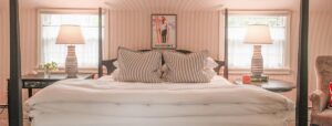 Bright bedroom with a large bed, two striped lamps on side tables, two windows with sheer curtains, a flower vase, an armchair, and an Edson Hill Guest Rooms Vermont poster above the bed.