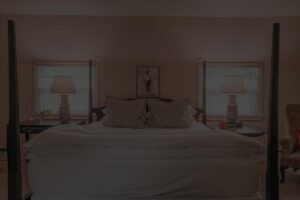 A cozy bedroom reminiscent of the Edson Hill Guest Rooms, featuring a large bed, two bedside tables with lamps, two windows with sheer curtains, and a framed Vermont poster on the back wall.