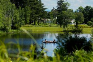 Two people relax on a small raft in the middle of a calm, tree-lined pond on a sunny day, with grassy land and houses visible in the background—a scene that perfectly captures the tranquil promise of vacation packages designed for peaceful getaways.