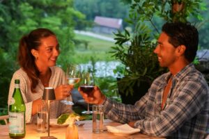 A woman and a man sit at an outdoor table, smiling and toasting with wine glasses as they enjoy a relaxing meal reminiscent of those included in luxury vacation packages. There are water glasses, a candle, and a bottle of sparkling water on the table, with greenery visible in the background.