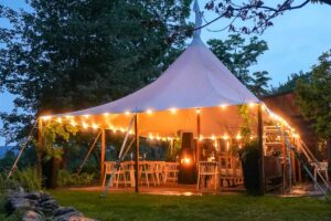 A white event tent is lit with string lights at dusk, surrounded by trees and greenery—offering the enchanting atmosphere often found in exclusive vacation packages. White chairs and tables are set up underneath for guests on a wooden platform, completing the picturesque scene.