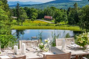 Elegant outdoor dining table with white flowers and candles overlooks a pond, green trees, a rustic barn, and distant mountains under a bright blue sky—the perfect setting for unforgettable moments often featured in luxury vacation packages.