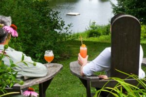 Two people sit in wooden chairs facing a lake, holding colorful drinks. Flowers and greenery surround them, and a small floating dock is visible on the water—an idyllic scene that captures the essence of relaxing vacation packages.