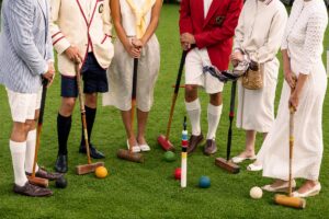 Six people dressed in vintage-style clothing stand in a circle on grass, holding croquet mallets—a scene reminiscent of classic leisure escapes. Croquet balls and a wicket are on the ground in the center of the group, evoking the timeless elegance often found in curated vacation packages.