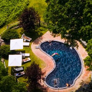 Aerial view of a kidney-shaped pool surrounded by grass and trees, with people swimming and lounging under white umbrellas on poolside chairs—an inviting scene perfect for those seeking unforgettable vacation packages.