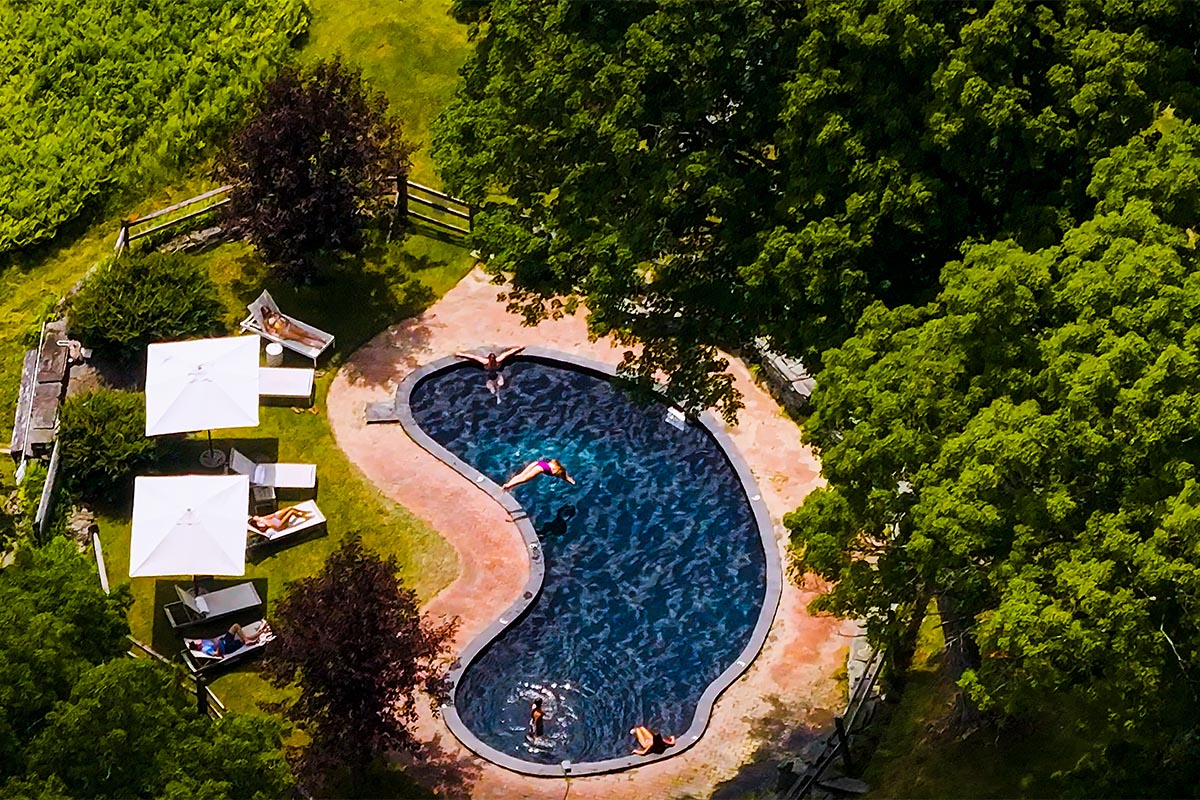 edson-hill-pool Aerial view of a kidney-shaped pool surrounded by grass and trees, with people swimming and lounging under white umbrellas on poolside chairs—an inviting scene perfect for those seeking unforgettable vacation packages.