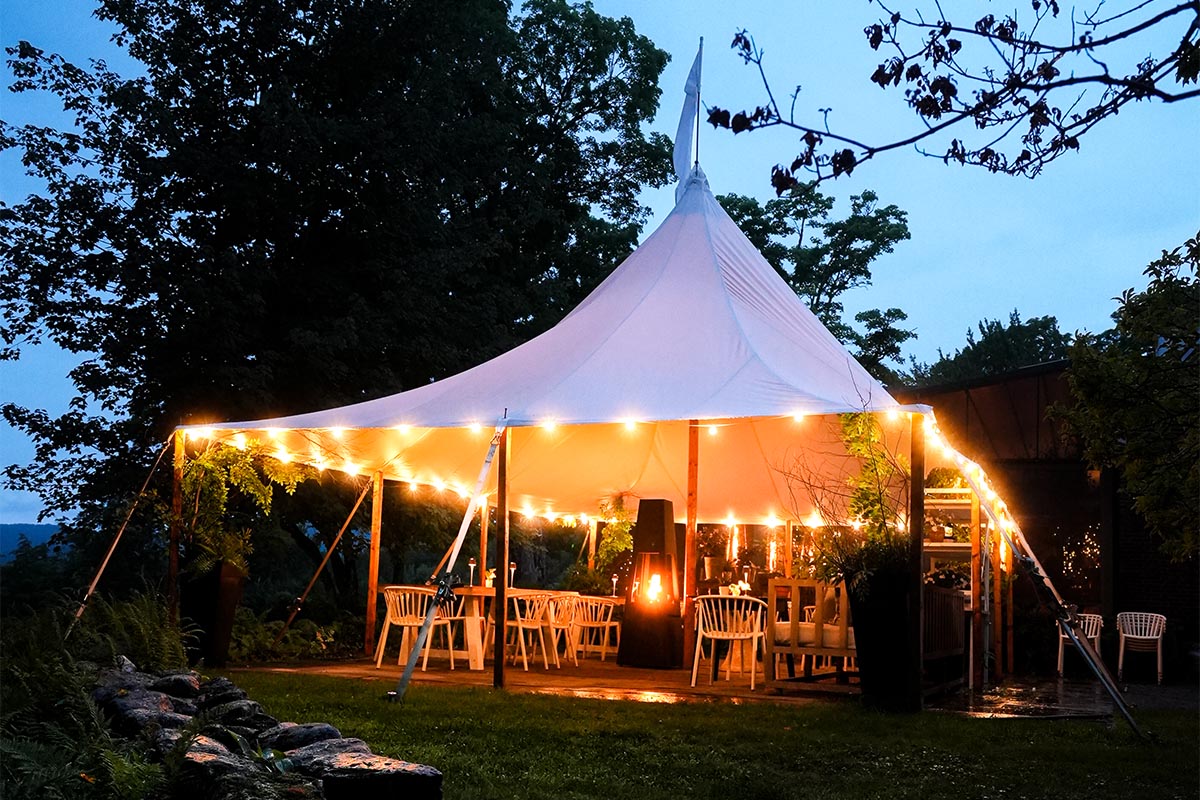 event-tent A white tent with string lights shelters tables and chairs in an outdoor setting at dusk, surrounded by trees and greenery. The atmosphere is warm and inviting.