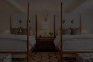 Two four-poster beds with white bedding and decorative pillows face a shared nightstand with a lamp, flowers, and antler wall decor in a cozy, symmetrical bedroom inspired by the refined charm of Edson Hill.
