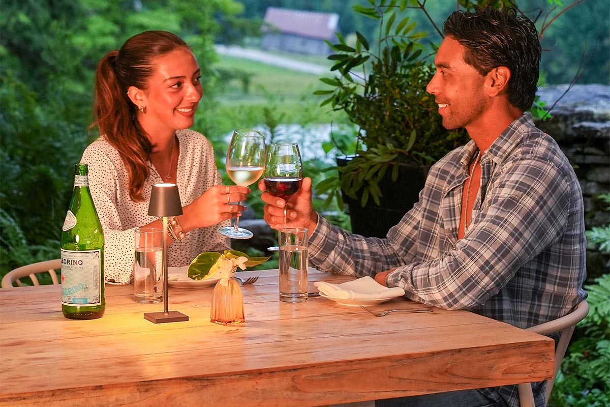 patio-exterior A woman and man sit at an outdoor table, smiling and toasting with wine glasses. They are surrounded by greenery and trees, with a bottle of sparkling water on the table.