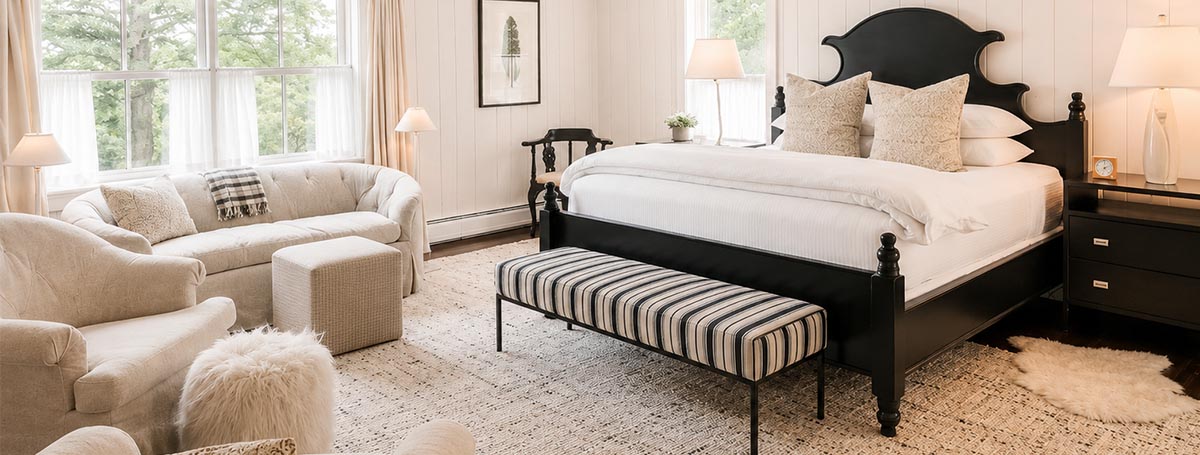 Bright bedroom with large windows at Edson Hill, a black framed bed, white bedding, striped bench, cozy white armchairs, a sofa, and soft lighting from table lamps. Neutral tones create an inviting, elegant atmosphere. Bright bedroom with large windows at Edson Hill, a black framed bed, white bedding, striped bench, cozy white armchairs, a sofa, and soft lighting from table lamps. Neutral tones create an inviting, elegant atmosphere.