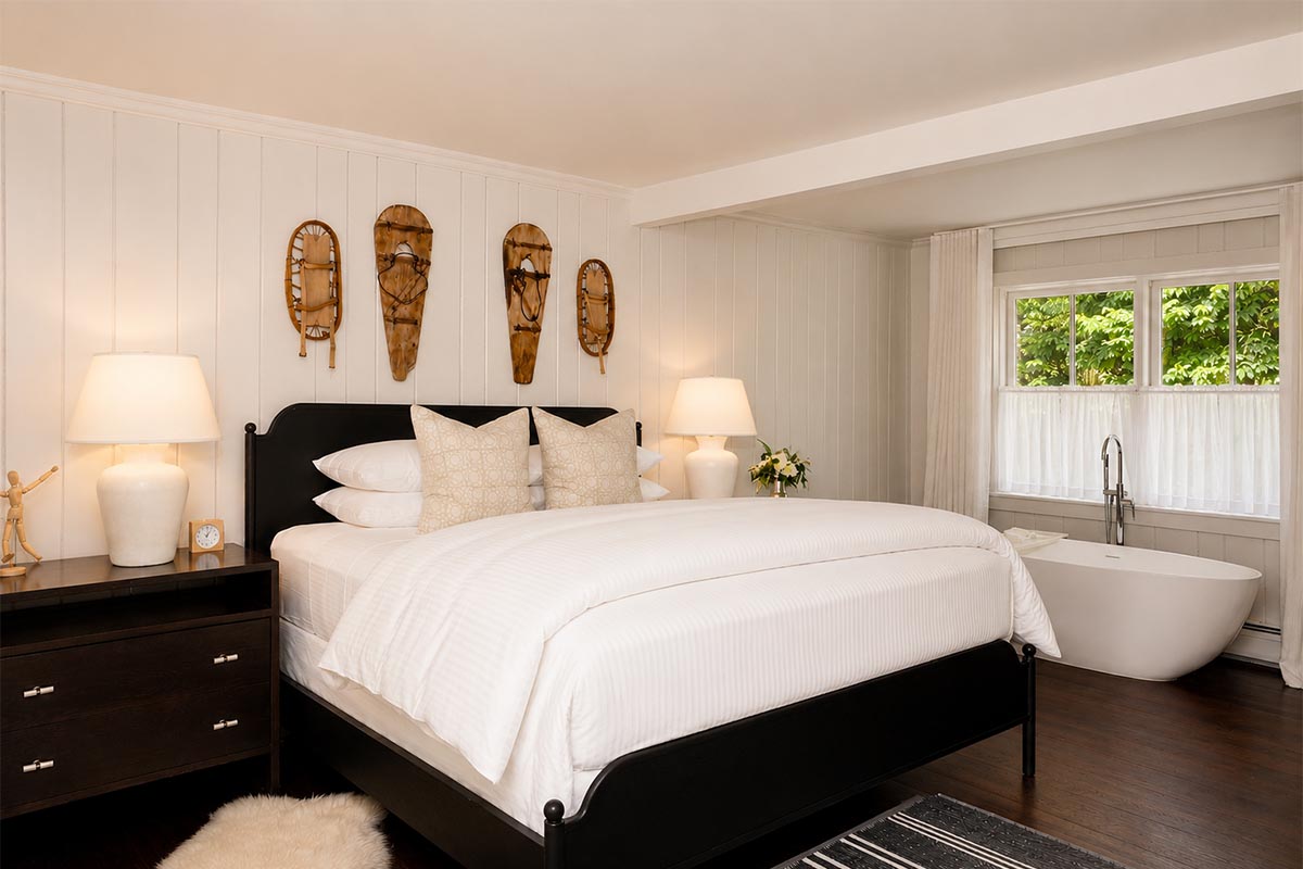 room-19 A cozy, modern bedroom—perfect for relaxing after exploring exciting vacation packages—features a black bed frame, white bedding, two beige pillows, nightstands with lamps, and three decorative wooden masks on the wall above the bed. A window lets in natural light.