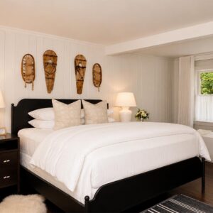 A cozy, modern bedroom—perfect for relaxing after exploring exciting vacation packages—features a black bed frame, white bedding, two beige pillows, nightstands with lamps, and three decorative wooden masks on the wall above the bed. A window lets in natural light.