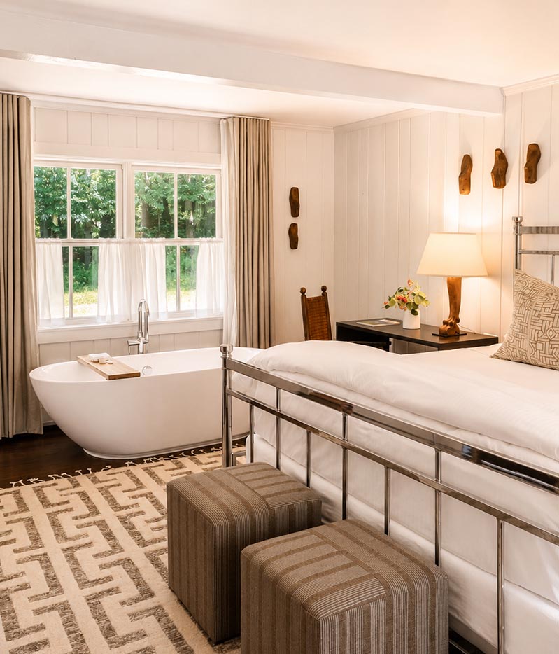 A cozy bedroom reminiscent of Vermont luxury hotel accommodations features a freestanding bathtub near large windows, a metal bed with white bedding, a patterned rug, two striped stools, and a nightstand with a lamp and flowers.