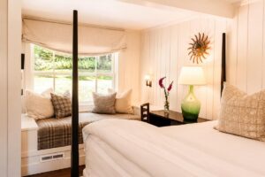 Cozy bedroom with a window seat, decorative pillows, a plaid throw, a dark wooden desk with a green lamp, and a sunburst wall decor above. Sunlight streams in through the large window, brightening the space—evoking the timeless charm of Edson Hill - Stowe Vermont Inn.