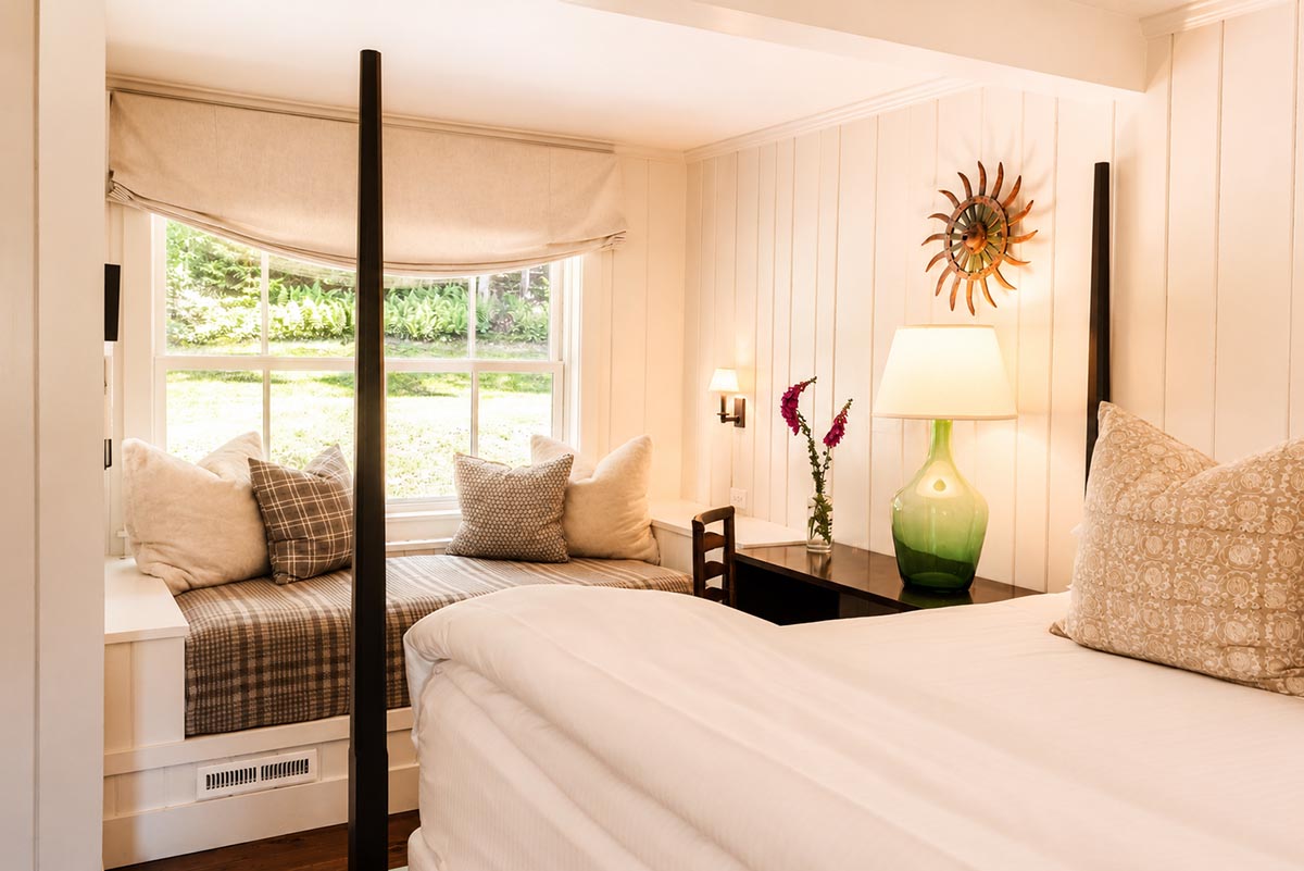 Cozy bedroom with a window seat, decorative pillows, a plaid throw, a dark wooden desk with a green lamp, and a sunburst wall decor above. Sunlight streams in through the large window, brightening the space—evoking the timeless charm of Edson Hill - Stowe Vermont Inn. Cozy bedroom with a window seat, decorative pillows, a plaid throw, a dark wooden desk with a green lamp, and a sunburst wall decor above. Sunlight streams in through the large window, brightening the space—evoking the timeless charm of Edson Hill - Stowe Vermont Inn.