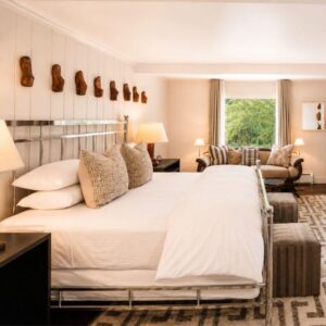 Bright hotel room with a large neatly made bed, decorative pillows, patterned rug, sofas by a window, and wall art featuring wooden sculptures above the headboard—an inviting retreat perfect for those seeking comfort and style as part of their vacation packages.