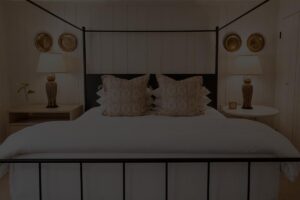 A neatly made bed with white linens, patterned pillows, and two owl-shaped lamps on bedside tables creates a cozy atmosphere in the Edson Hill Guest Rooms. Decorative plates hang on the white paneled wall behind the bed, adding a charming touch to this inviting space.