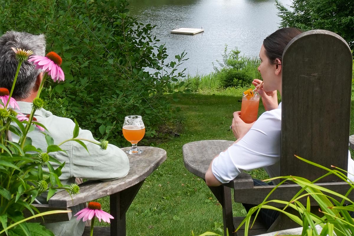 summer-drinks-chair Two people sit in wooden chairs by a small lake, holding colorful drinks. They are surrounded by green grass, trees, and pink flowers, with a dock floating on the water in the distance.