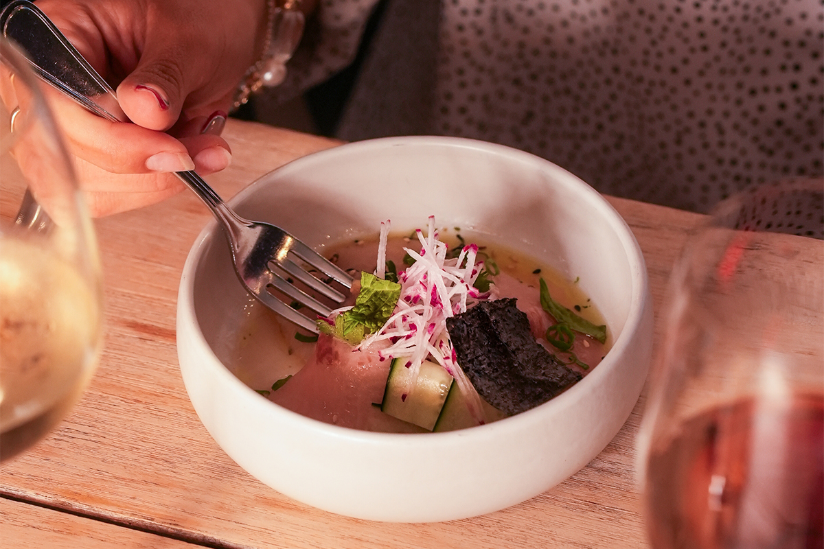 A person holds a fork over a bowl of soup garnished with sliced radish, cucumber, herbs, and a piece of black seaweed, with two glasses of wine visible on the wooden table—a delightful scene reminiscent of culinary experiences found in curated vacation packages. A person holds a fork over a bowl of soup garnished with sliced radish, cucumber, herbs, and a piece of black seaweed, with two glasses of wine visible on the wooden table—a delightful scene reminiscent of culinary experiences found in curated vacation packages.