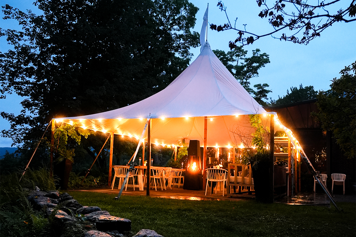 A white tent with string lights glows at dusk, sheltering white chairs and tables on a patio—perfect for guests enjoying vacation packages. Trees and greenery surround the cozy outdoor setting. A white tent with string lights glows at dusk, sheltering white chairs and tables on a patio—perfect for guests enjoying vacation packages. Trees and greenery surround the cozy outdoor setting.