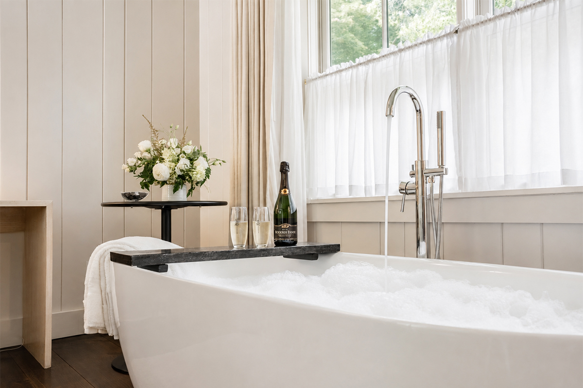 A modern bathroom with a white freestanding tub filled with bubbles, a champagne bottle and glasses on a tray, white flowers on a side table, and sheer curtains covering tall windows—perfect for unwinding after exploring the best vacation packages.